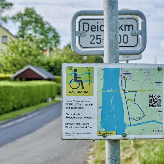 Nahaufnahme einer Wegbeschilderung für einen rollstuhlgeeigneten Rundweg in Loxstedt. Im Hintergrund sind ein Deich, ein geteerter Weg und Häuser zu sehen