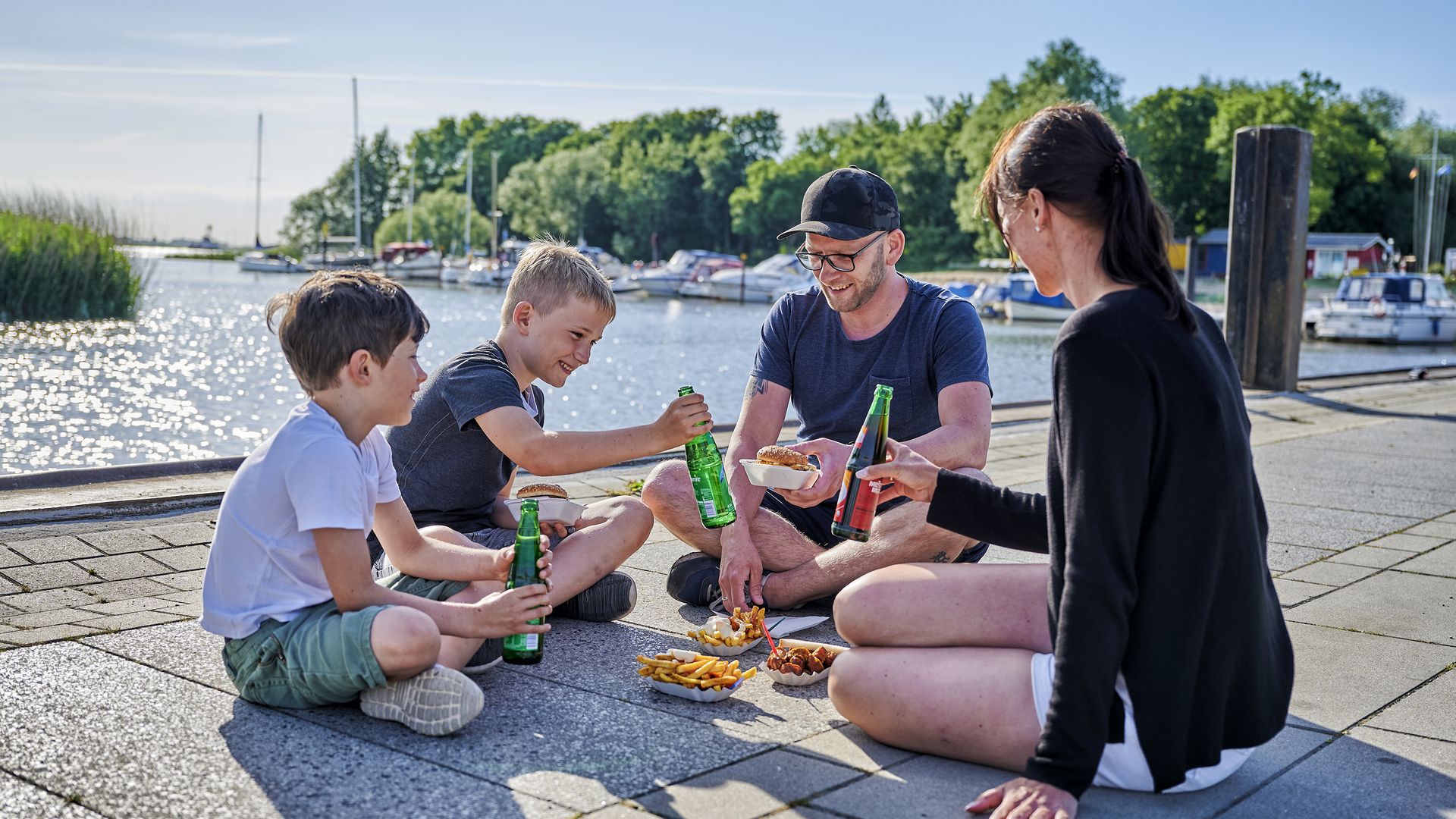 Am Hafenbecken von Sandstedt sitzt eine vierköpfige Familie bei strahlendem Sonnenschein auf dem Boden, isst Pommes und Currywurst und trinkt Limonade. Im Hintergrund sind Boote zu sehen, die im Hafen festgemacht haben. 