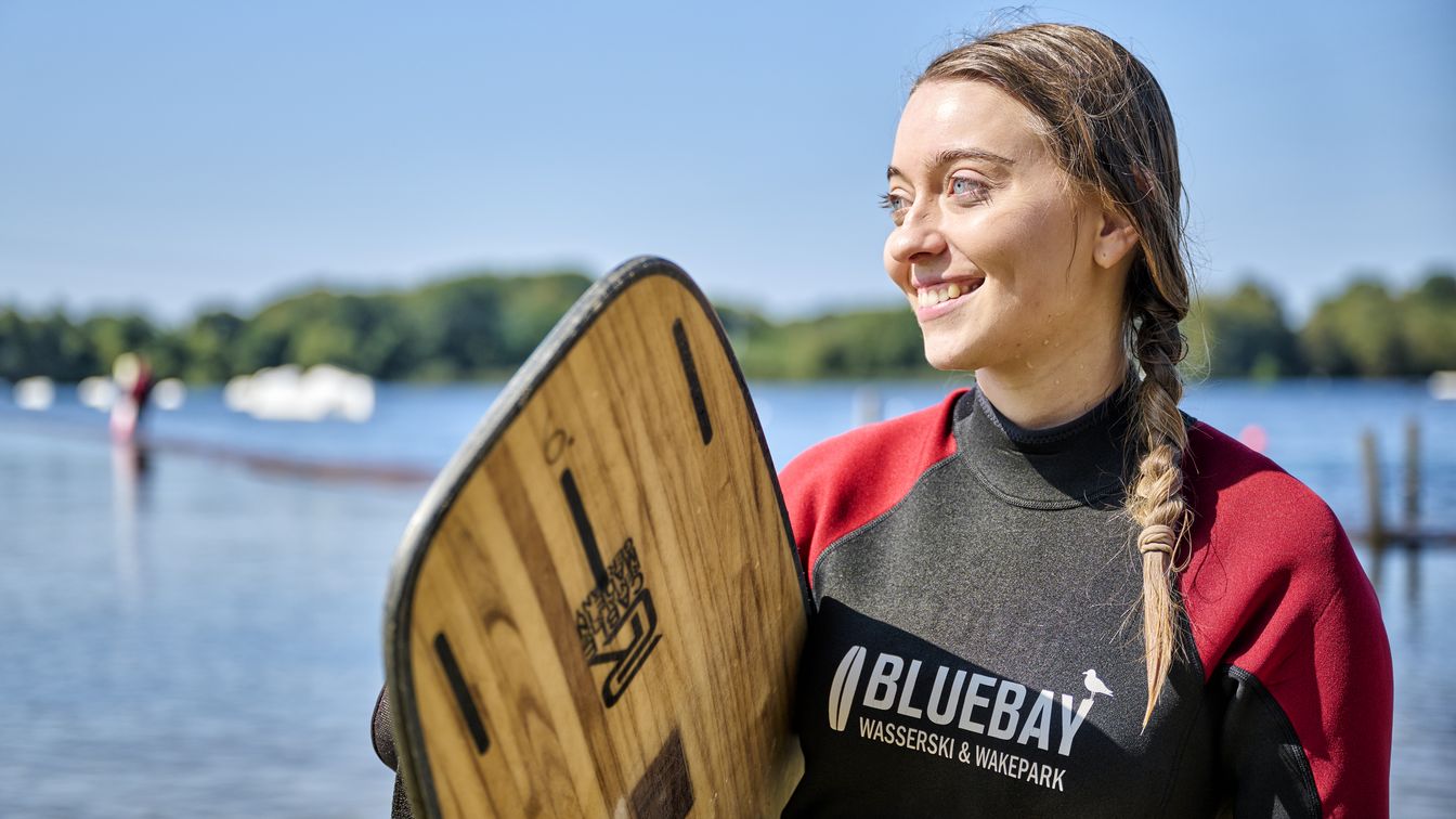 Portraitaufnahme einer lächelnden, blonden Wakeboarderin, die ihr Wakeboard unter dem Arm trägt. Sie hat einen rot-schwarzen Neoprenanzug an. Sie hat blaue Augen und die Harre zu einem Zopf geflochten, der ihr auf die rechte Schulter fällt.