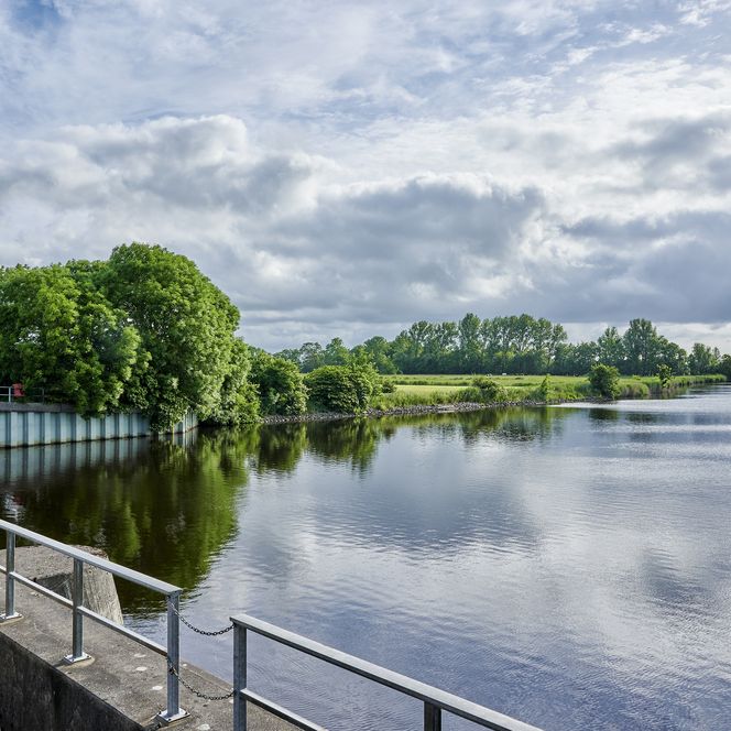 Blick von der Schleuse aus auf einen Fluss, die linke Uferseite und den angrenzenden Weiden.