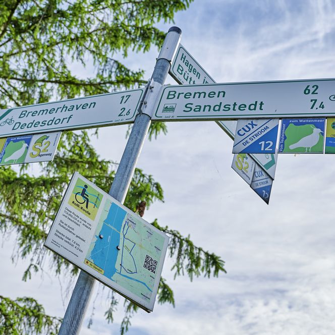 Wegbeschilderung für einen rollstuhlgeeigneten Rundweg in Loxstedt. Im Hintergrund ist ein grüner Baum und Wolkenhimmel zu sehen.