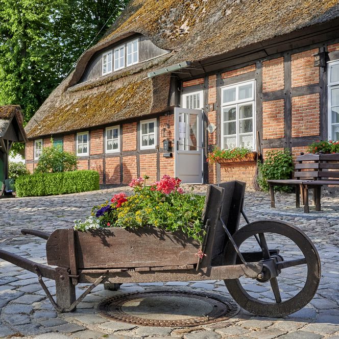 Blick auf den Eingangsbereich des Niedersachsenhauses in Bramstedt. Auf dem Vorplatz aus Kopfsteinpflastern steht eine alte Schubkarre aus Holz, die mit Blumen bepflanzt ist. Dahinter sieht man eine runde Bank, die vor dem Haus steht. 