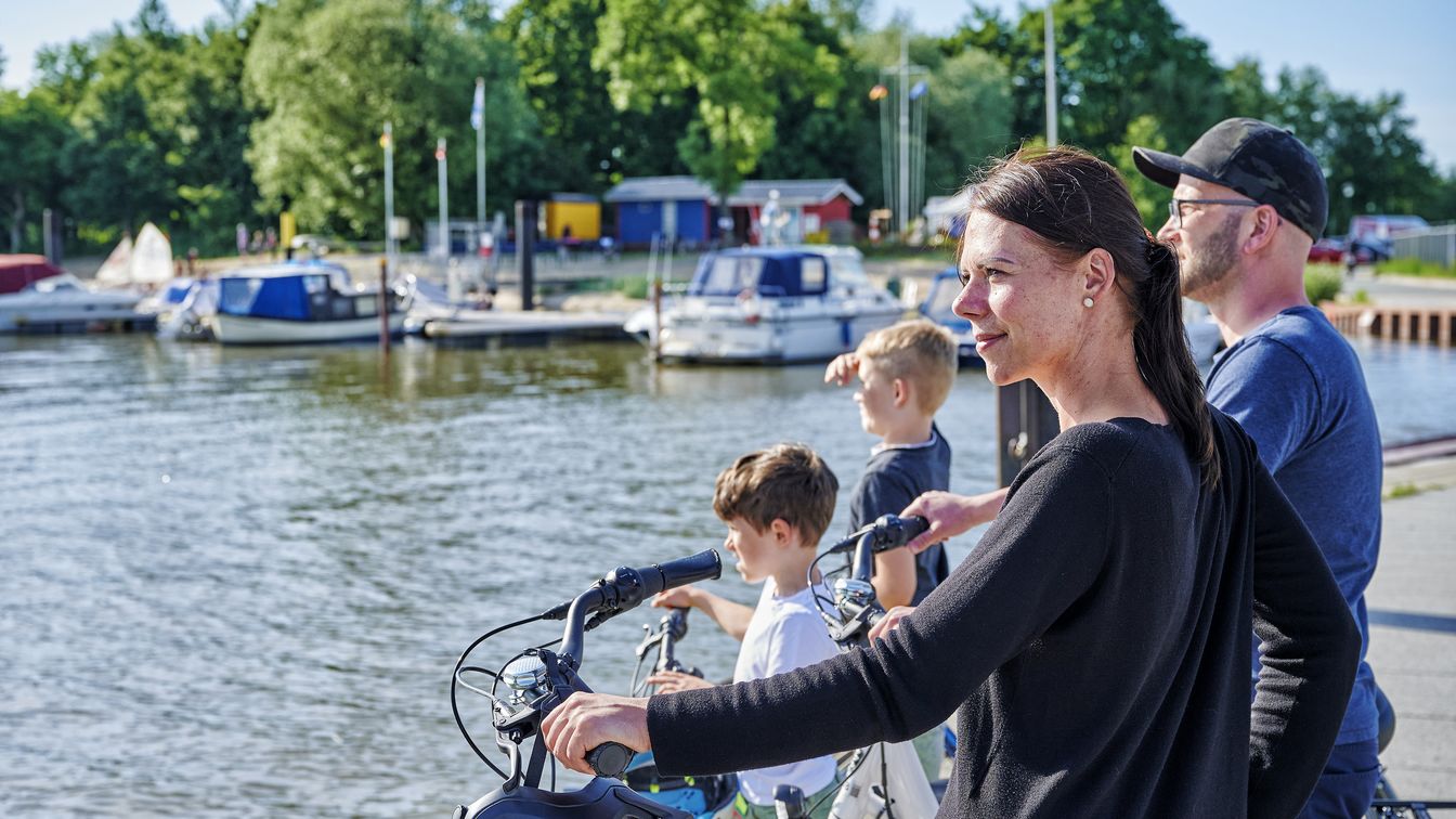 Familie mit zwei kleinen Kindern, die mit ihren Fahrrädern an der Pier stehen und auf das Wasser schauen. Das eine Kind hält sich die Hand vor die Stirn als Sonnenschutz. Im Hintergrund sieht man eine Anlegestelle mit vielen kleinen Booten und Häuschen. Die Sonne scheint.